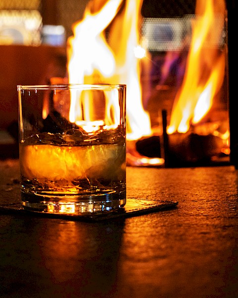 A glass of whiskey or similar drink sits on a coaster in front of a warm, glowing fire, creating a cozy atmosphere.