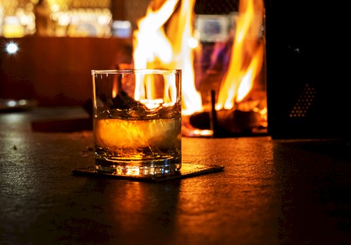 A glass of whiskey or similar drink sits on a coaster in front of a warm, glowing fire, creating a cozy atmosphere.