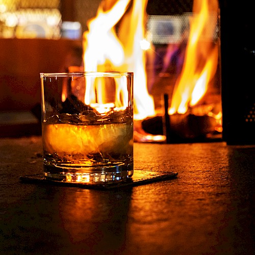 A glass of whiskey or similar drink sits on a coaster in front of a warm, glowing fire, creating a cozy atmosphere.