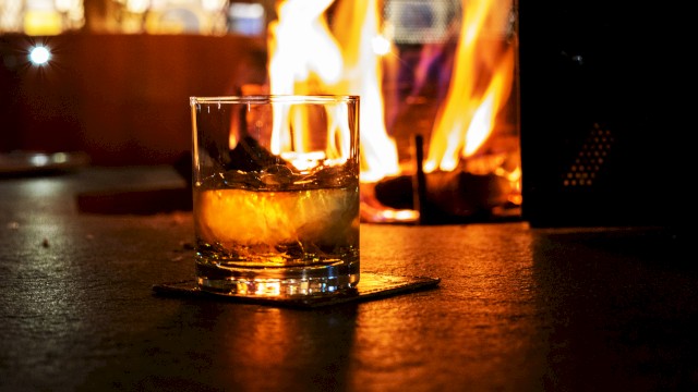 A glass of whiskey or similar drink sits on a coaster in front of a warm, glowing fire, creating a cozy atmosphere.