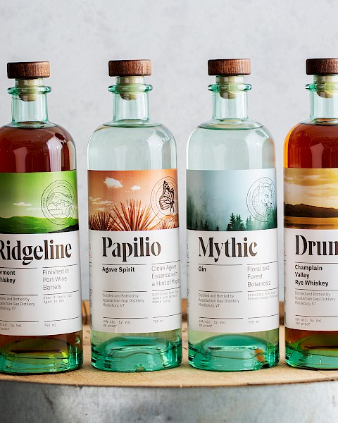 Four bottles of spirits labeled Ridgeline, Papilio, Mythic, and Drumlin, displayed on a wooden barrel.