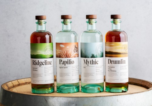 Four bottles of spirits labeled Ridgeline, Papilio, Mythic, and Drumlin, displayed on a wooden barrel.
