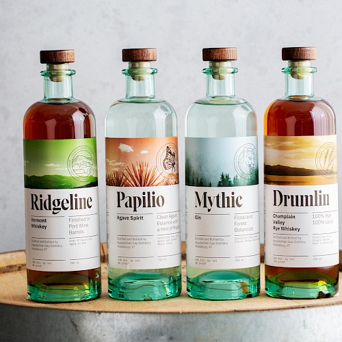 Four bottles of spirits labeled Ridgeline, Papilio, Mythic, and Drumlin, displayed on a wooden barrel.