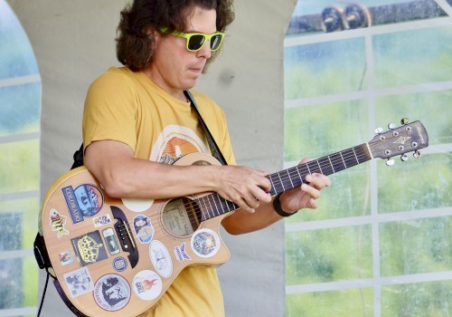A person in sunglasses plays a guitar decorated with stickers, wearing a yellow shirt and blue plaid shorts, under a tent.