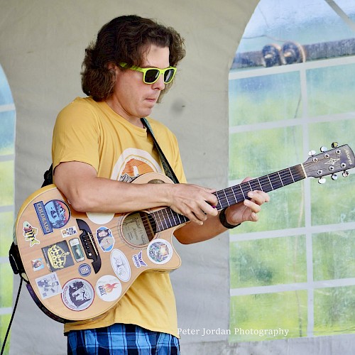 A person in sunglasses plays a guitar decorated with stickers, wearing a yellow shirt and blue plaid shorts, under a tent.