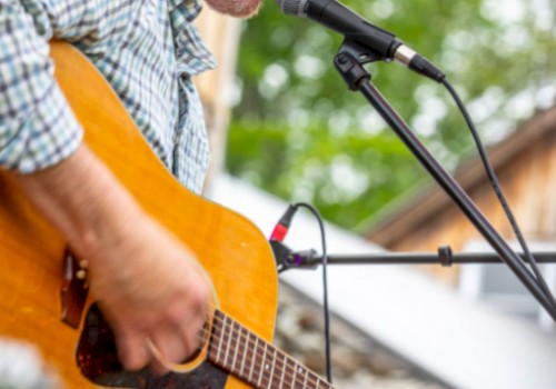 A man is playing an acoustic guitar and singing into a microphone outdoors, wearing a cap and a plaid shirt.