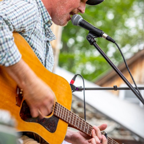 A man is playing an acoustic guitar and singing into a microphone outdoors, wearing a cap and a plaid shirt.