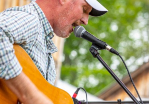 A person is playing guitar and singing into a microphone outdoors, wearing a cap and plaid shirt.