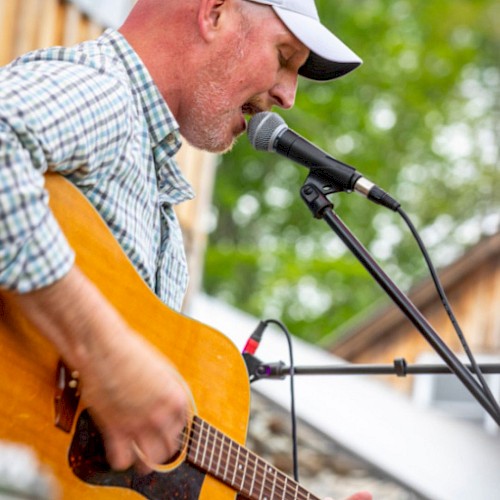 A person is playing guitar and singing into a microphone outdoors, wearing a cap and plaid shirt.