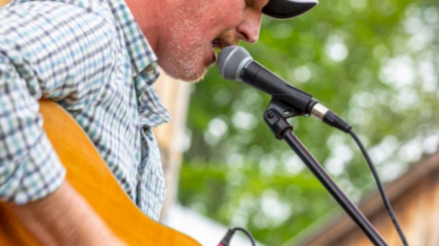 A person is playing guitar and singing into a microphone outdoors, wearing a cap and plaid shirt.