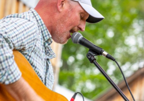 A person is playing an acoustic guitar and singing into a microphone outdoors, wearing a checked shirt and a cap.