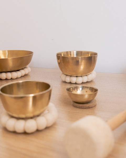A close-up of several brass singing bowls arranged on a wooden surface, each resting on soft white felt rings. A padded wooden mallet lies nearby, evoking a sense of calm and mindfulness often associated with sound therapy or meditation practices.