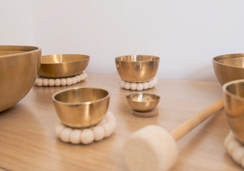 A close-up of several brass singing bowls arranged on a wooden surface, each resting on soft white felt rings. A padded wooden mallet lies nearby, evoking a sense of calm and mindfulness often associated with sound therapy or meditation practices.