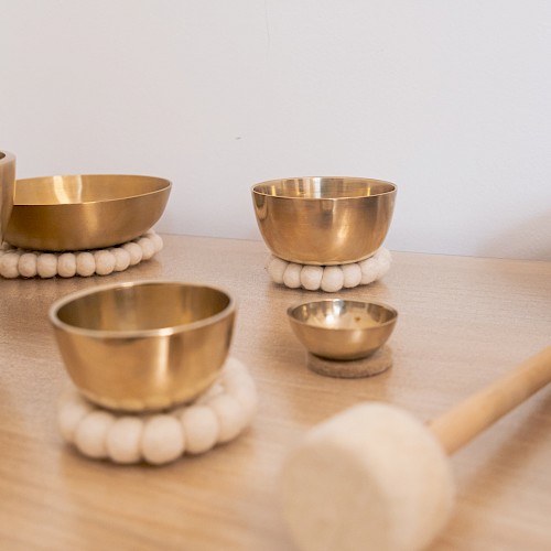 A close-up of several brass singing bowls arranged on a wooden surface, each resting on soft white felt rings. A padded wooden mallet lies nearby, evoking a sense of calm and mindfulness often associated with sound therapy or meditation practices.