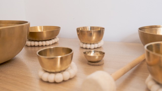 A close-up of several brass singing bowls arranged on a wooden surface, each resting on soft white felt rings. A padded wooden mallet lies nearby, evoking a sense of calm and mindfulness often associated with sound therapy or meditation practices.