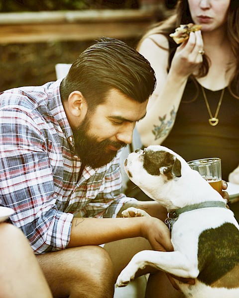 A man interacts with a dog at a casual gathering, while others enjoy food and drinks, creating a warm, relaxed atmosphere.
