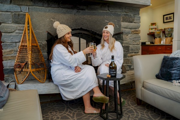 Two people in robes and beanies toast drinks by a fireplace, with snowshoes on the wall and a bottle on a small table.