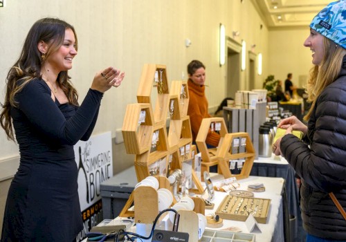 A woman in black smiles while showing jewelry to another woman at a market stall with wooden displays and various items for sale.