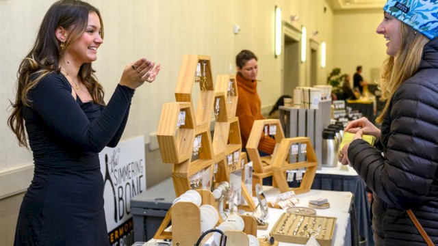 A woman in black smiles while showing jewelry to another woman at a market stall with wooden displays and various items for sale.