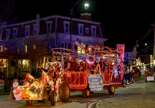 A festive parade float decorated with holiday lights and characters moves through a street at night, featuring a Christmas theme.