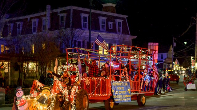 A festive parade float decorated with holiday lights and characters moves through a street at night, featuring a Christmas theme.