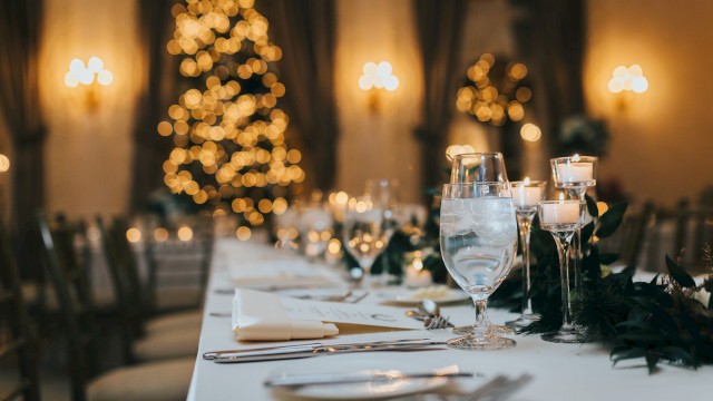 A festive table is set with glassware, candles, and greenery; a blurred, lit Christmas tree stands in the softly lit background.