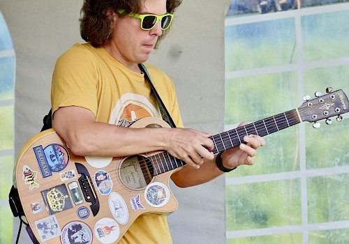 A person with sunglasses plays a sticker-covered guitar at an outdoor event, wearing a yellow shirt and blue checkered shorts.