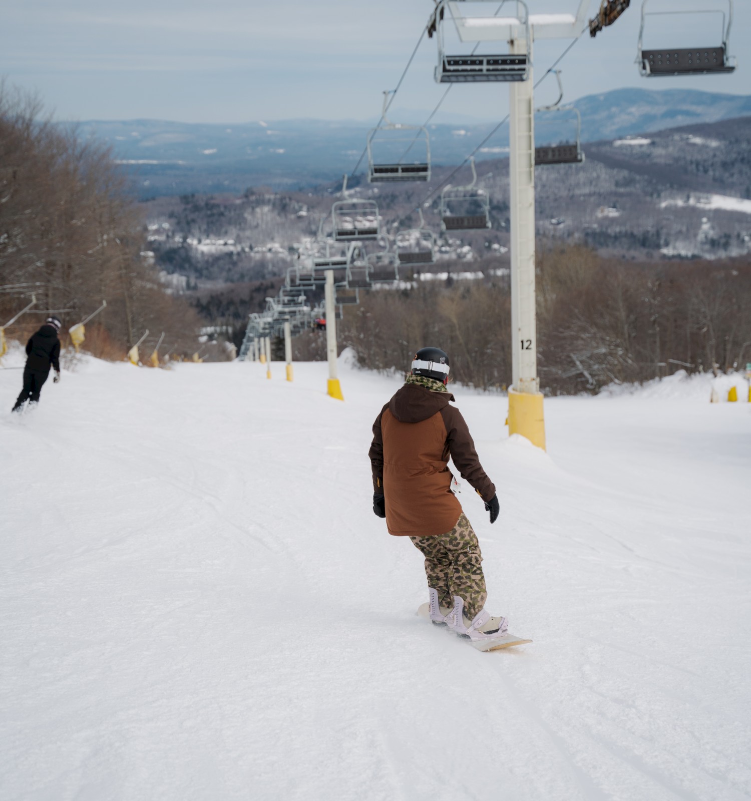 A person snowboarding down a snowy slope beneath a ski lift, with a scenic winter landscape in the background.