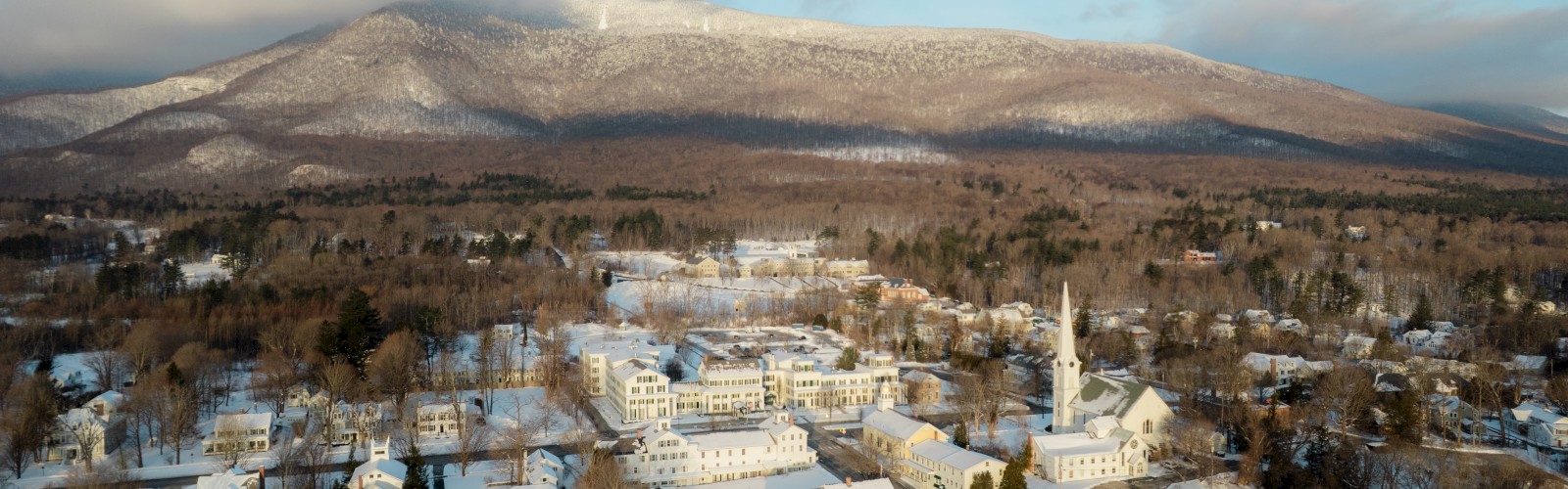 A snowy town is set against a backdrop of mountains, featuring several buildings and a tall white church steeple under a cloudy sky.