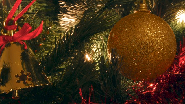 A festive Christmas tree close‑up with a glittery gold bauble, red ribbons, and twinkling lights among green needles, creating a cozy holiday glow.