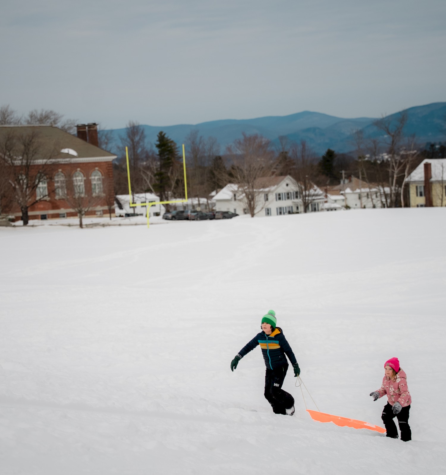 Two kids sledding in a snowy field near a small town; houses and trees line the background with mountains in the distance.