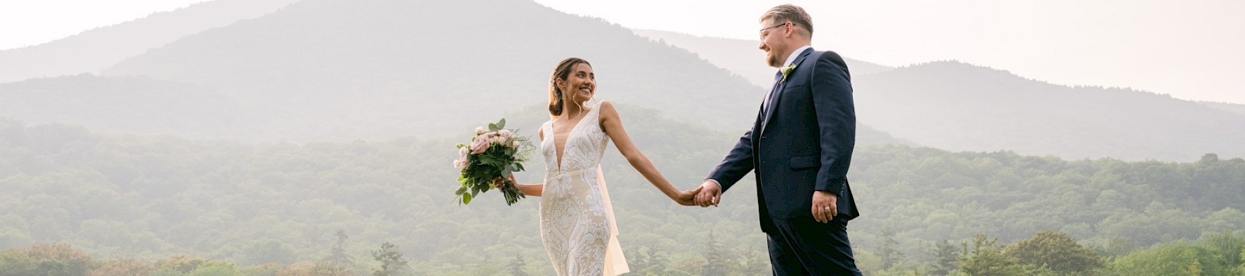 A newlywed couple holds hands on a hilltop, the bride in a lace dress with a bouquet, the groom in a dark suit, mountain scenery behind them.