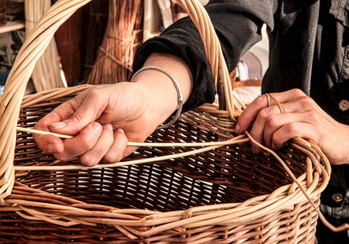 A close-up of hands weaving a wicker basket, with strands of reed and a handle visible, in a craft setup.