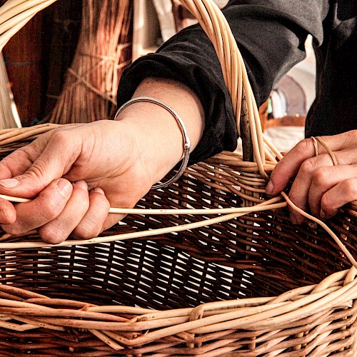 A close-up of hands weaving a wicker basket, with strands of reed and a handle visible, in a craft setup.