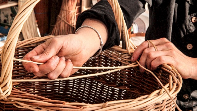 A close-up of hands weaving a wicker basket, with strands of reed and a handle visible, in a craft setup.