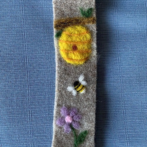 A felt bookmark featuring a sunny yellow sun, a buzzing bee, and purple flowers on a gray strip with a small green leaf near the top, end.