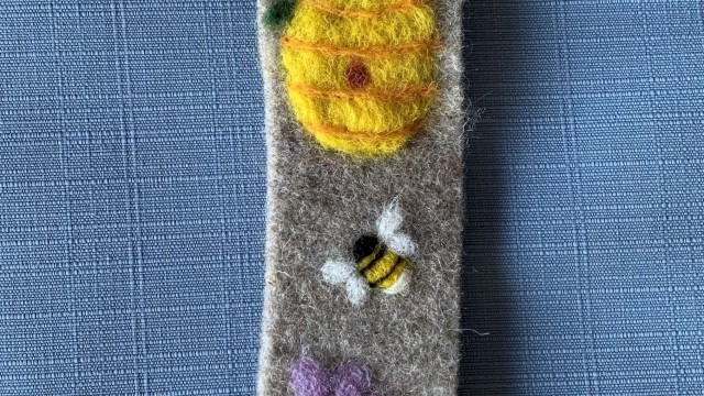 A felt bookmark featuring a sunny yellow sun, a buzzing bee, and purple flowers on a gray strip with a small green leaf near the top, end.