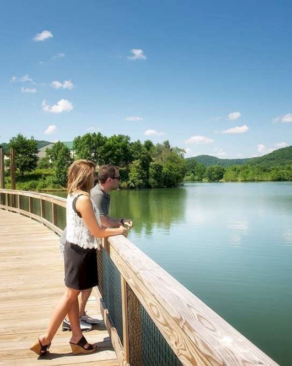 A couple stands on a wooden boardwalk overlooking a serene lake surrounded by lush green hills and a clear blue sky.