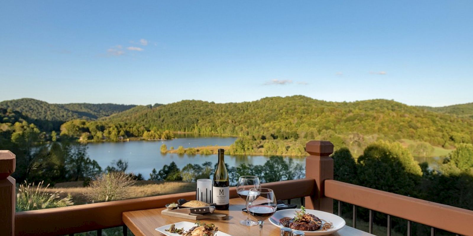 A picturesque outdoor dining scene with plates of food, wine, and glasses on a table overlooking a serene lake and forested hills.