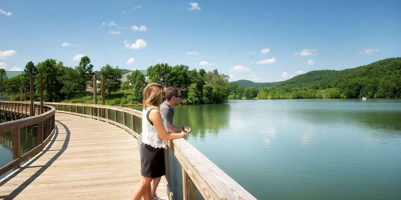 A couple stands on a wooden boardwalk overlooking a calm lake, surrounded by lush green hills and a clear blue sky.
