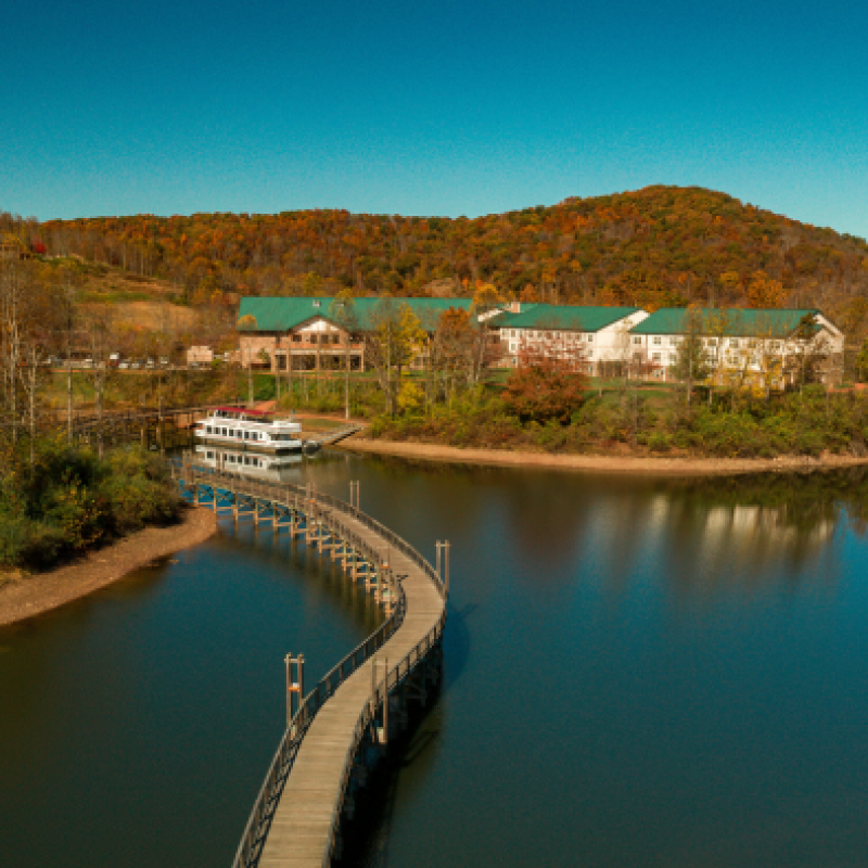 A scenic view of a lake with a winding bridge, surrounded by autumn trees and hills, leading to buildings with green roofs.