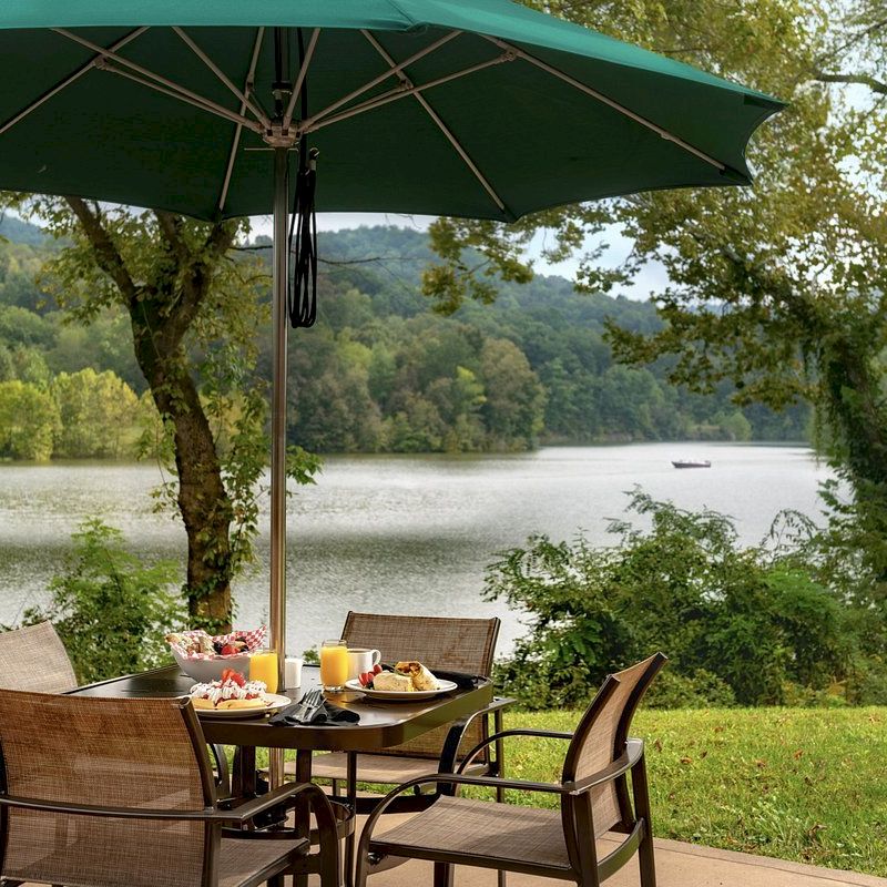 Outdoor dining setup with a table, chairs, and umbrella, overlooking a scenic lake surrounded by trees and hills in the background.