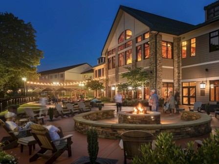 The image shows a cozy outdoor space with a fire pit, seating, and lights strung overhead, adjacent to a large building at dusk.