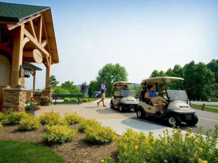A scene with two golf carts near a wooden building, lush greenery, and people carrying golf bags, all under a clear sky.