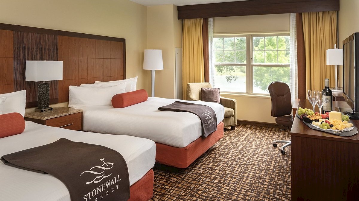 A hotel room with two beds, a window, lamp, armchair, and a dresser with wine and fruit, featuring 