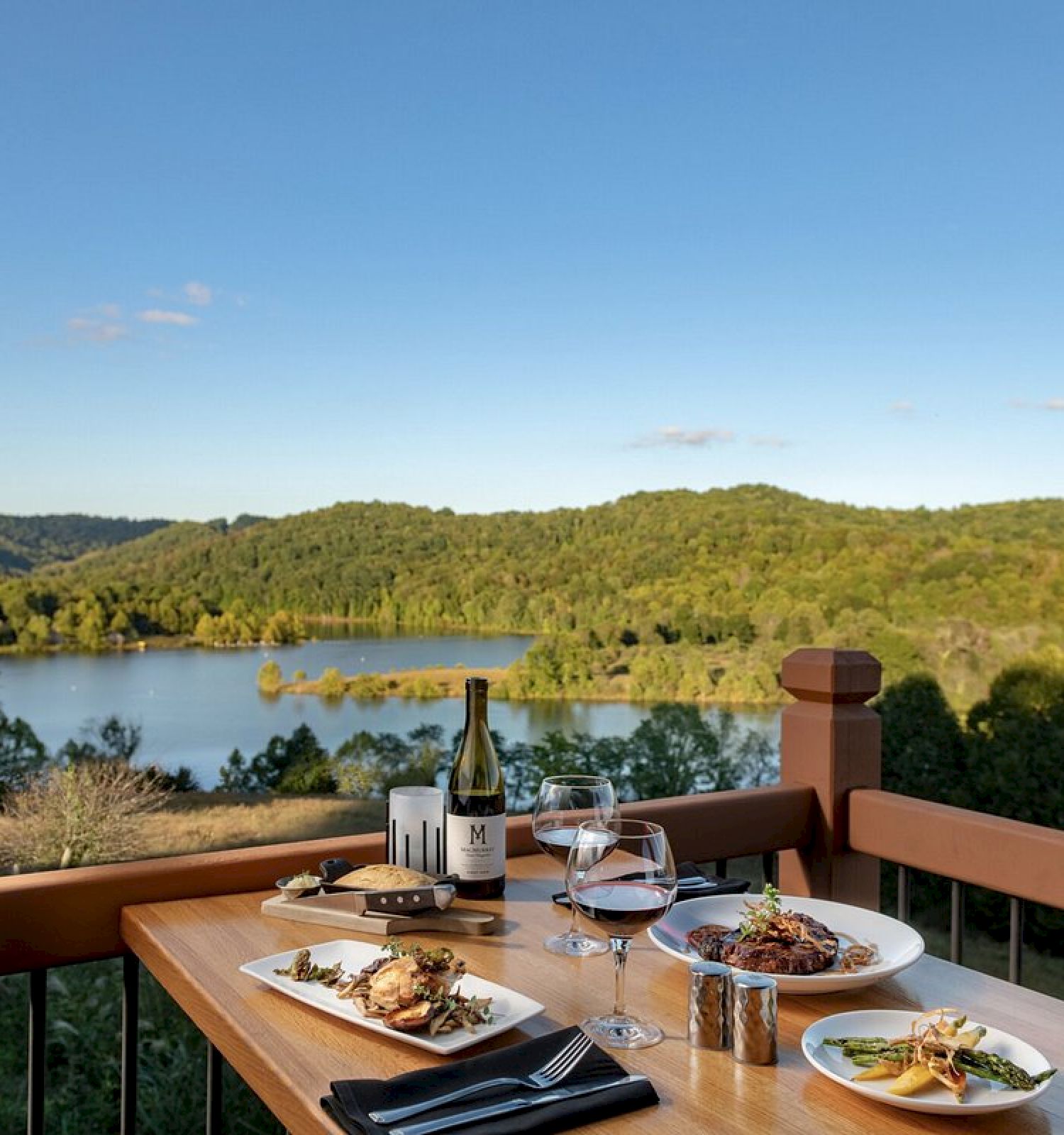 Outdoor dining setup with wine and food on a table overlooking a scenic lake and lush hills under a clear blue sky.