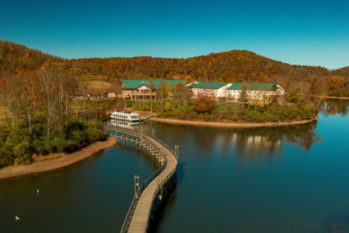 A wooden pathway curves over a lake leading to buildings surrounded by autumn trees and hills under a clear blue sky.