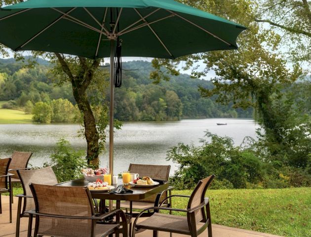 A patio table with food and drinks is under a green umbrella, overlooking a scenic lake and trees, with a peaceful boat in the distance.