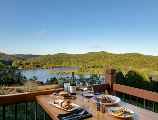 A dining table with plates of food and wine overlooks a scenic landscape with a lake and forested hills under a clear blue sky.