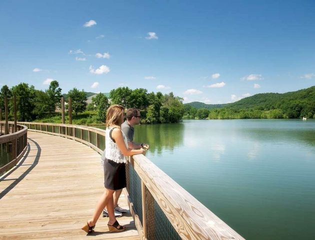 Two people stand on a wooden boardwalk overlooking a calm lake, surrounded by lush green hills under a clear blue sky.